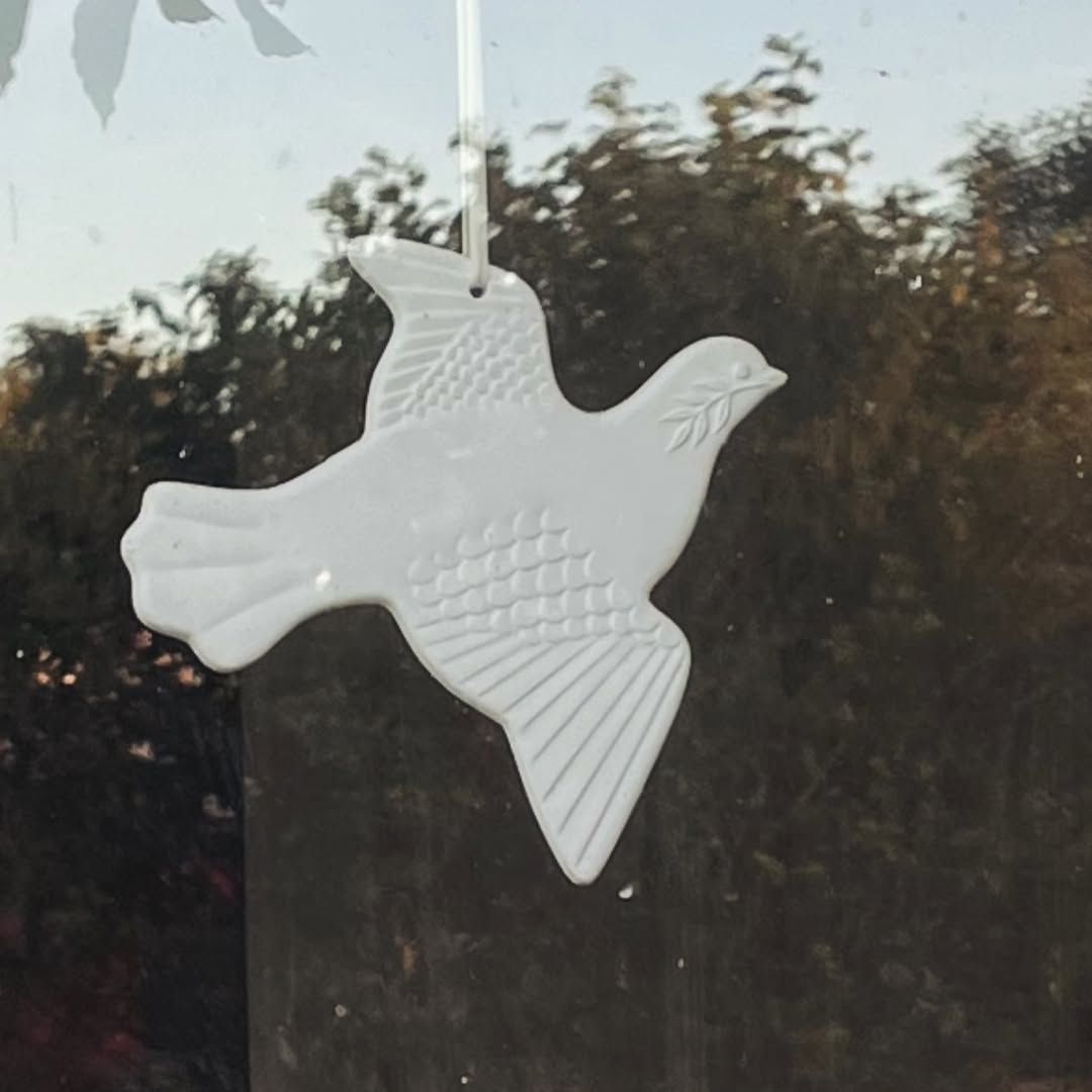 Biały ceramiczny gołąb zawieszony w oknie, na tle drzew